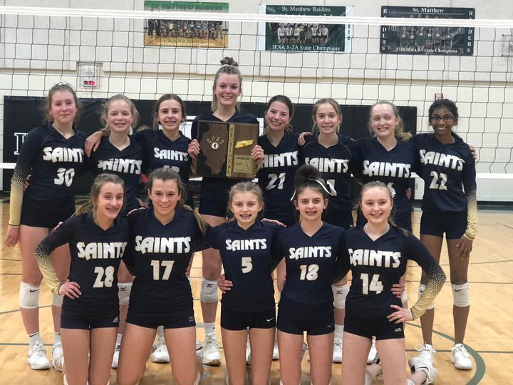 saints girls volleyball team posing with trophy in front of volleyball net