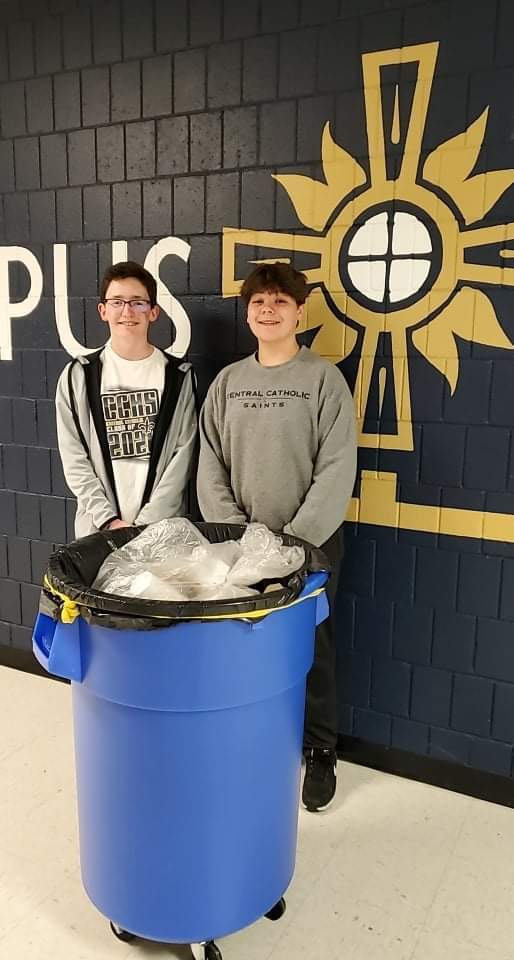 two boys smiling and standing behind blue garbage bin 